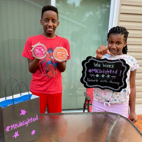 Two children holding delighted by dessert tubs beside a hand-lettered #MKdelighted sign