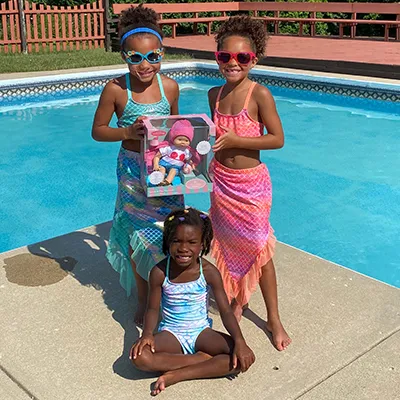 Three girls in mermaid swimsuits by a pool with a mermaid doll in its box
