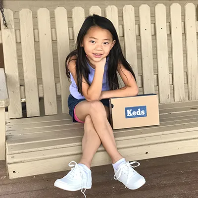 A girl on a porch swing with a Keds shoebox, wearing new white Keds